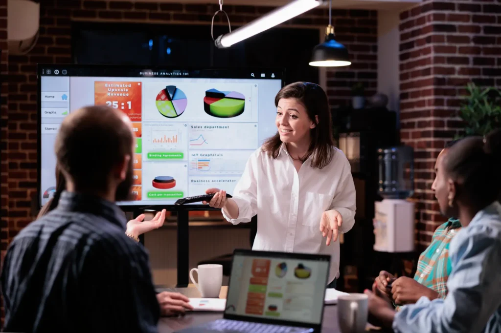 Smiling caucasian woman startup office doing business presentation big tv screen with charts front team confident employee presenting growing sales statistics late night meeting
